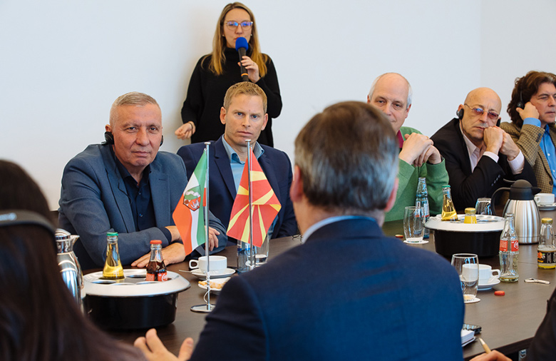 Eine Person spricht stehend in ein Mikrophon. Mehrere Teilnehmende sitzen in einem Tisch, auf dem eine NRW- und nordmazedonische Tischflagge zu sehen ist; eine Person ist von hinten zu sehen.